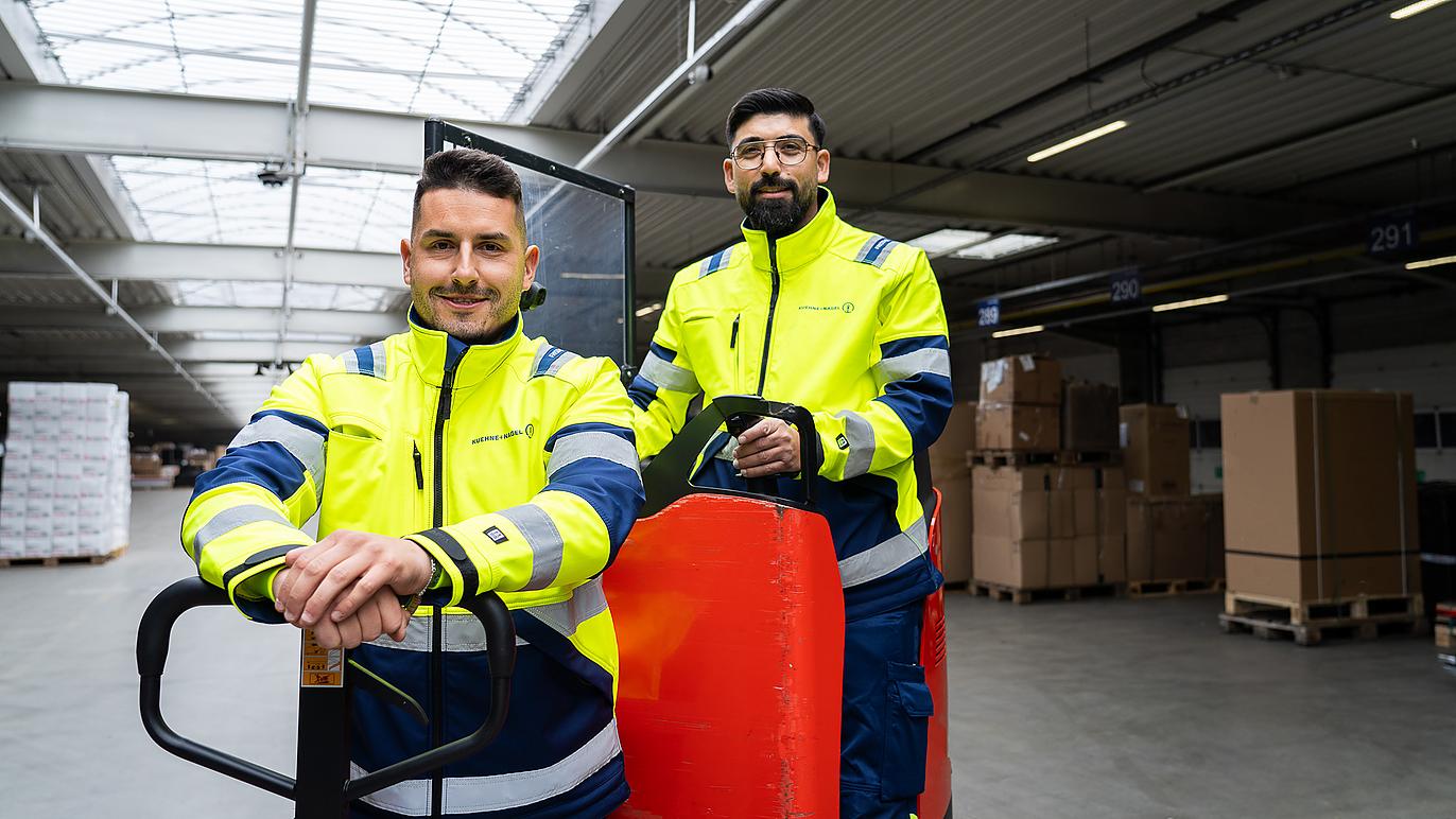 In einer hellen Lagerhalle posieren zwei Männer in Arbeitskleidung gut gelaunt auf einem Hubwagen.